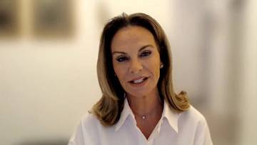 Kobieta o blond włosach, ubrana w białą koszulę, m&oacute;wi patrząc w kamerę.