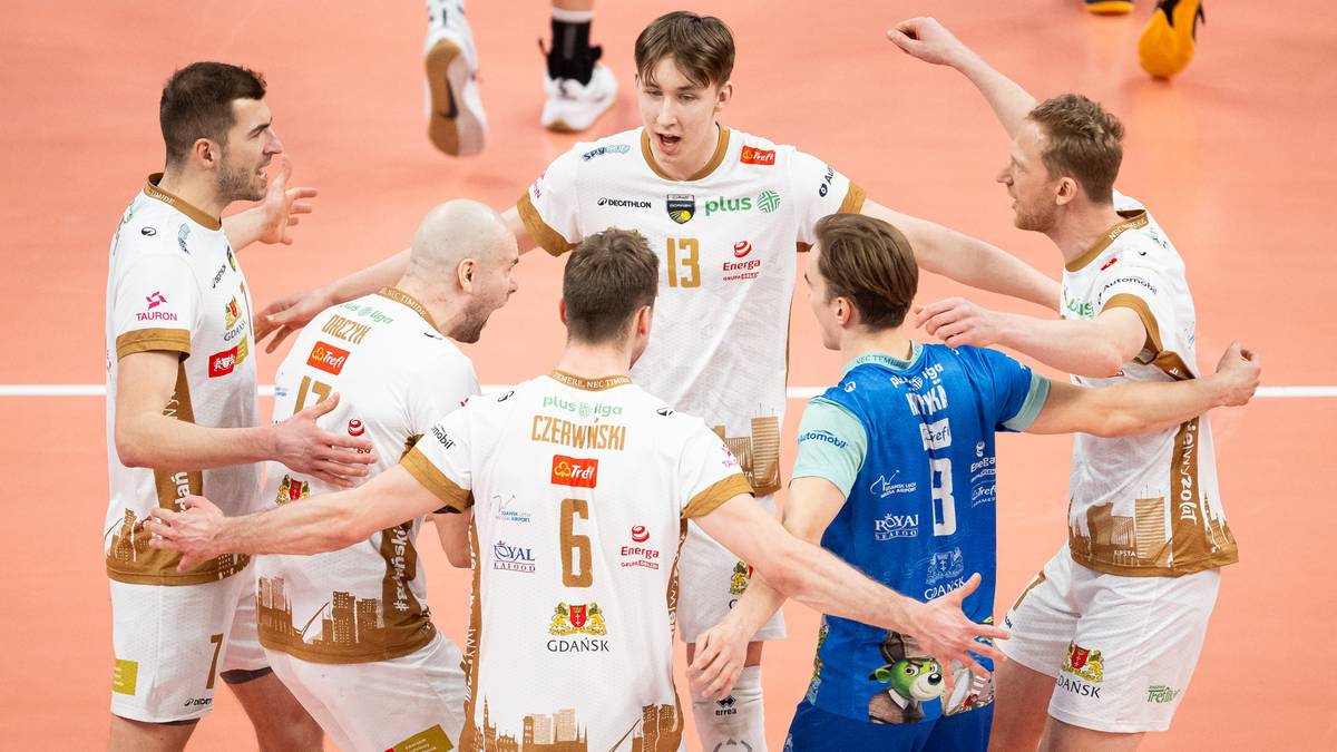 Grupa siatkarzy w białych strojach z ż&oacute;łtymi detalami świętuje zwycięstwo, przytulając się do siebie na parkiecie.