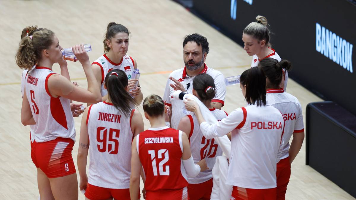 Grupa siatkarek w białych strojach z czerwonymi elementami i trener w otoczeniu dyskutuje na boisku.