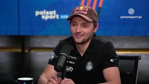 Andrzej Bargiel w czapce z daszkiem i czarnym polo siedzi przed mikrofonem, uśmiecha się.