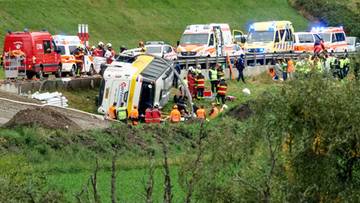 Przewr&oacute;cony na bok autobus na poboczu drogi oraz liczne pojazdy służb ratunkowych i ratownicy.