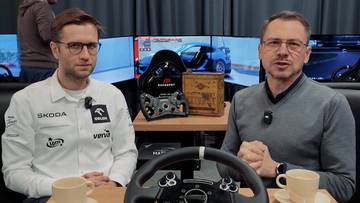 Dw&oacute;ch mężczyzn siedzi przy stole z kierownicą symulatora wyścigowego, na ekranach w tle widać gry wyścigowe.