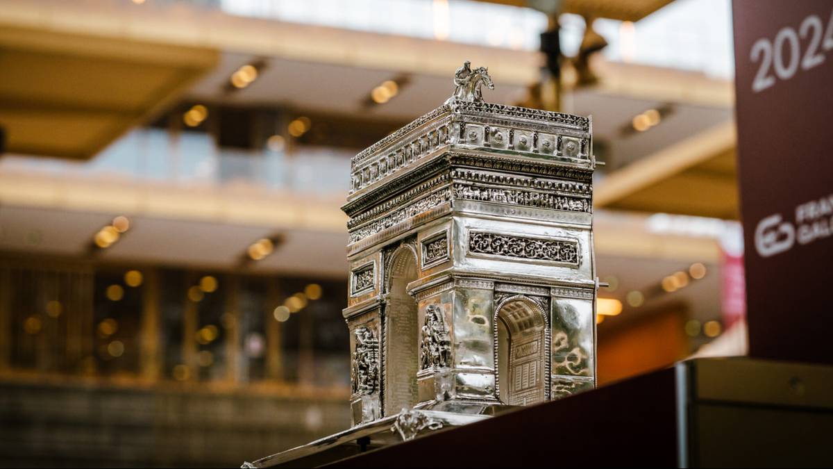 Miniaturowa replika Łuku Triumfalnego wykonana z metalu, ozdobiona detalami architektonicznymi, postawiona na cokole.