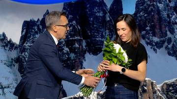 Mężczyzna w garniturze wręcza kobiecie bukiet białych kwiat&oacute;w.