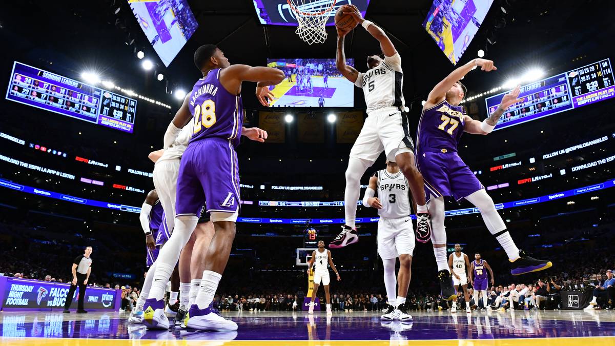 Koszykarze w akcji podczas meczu, jeden zawodnik wyskakuje do wsadu, inny próbuje go zablokować. Na drugim planie widać kibiców i parkiet.