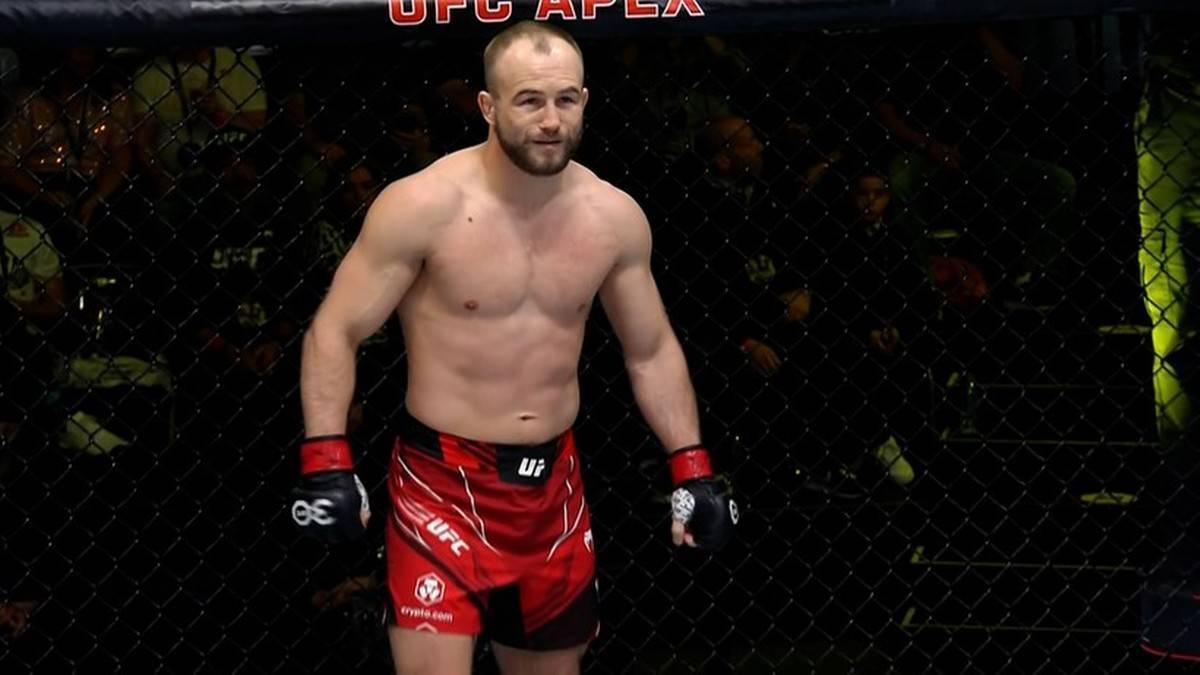 Zawodnik MMA w czerwonych spodenkach i rękawicach treningowych stoi w klatce.