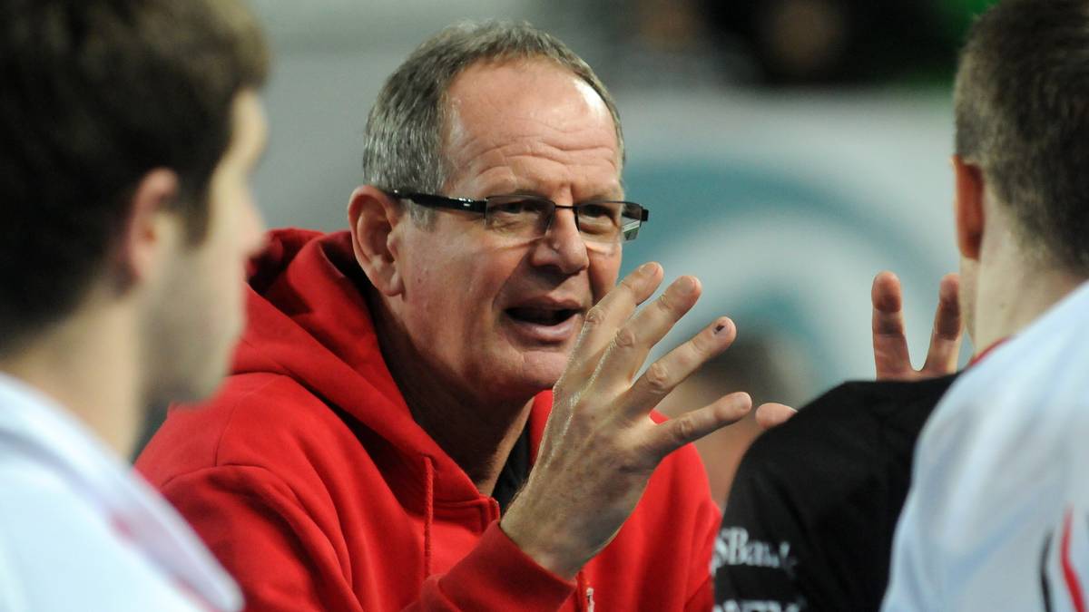 Starszy mężczyzna w okularach i czerwonej bluzie z kapturem, mówiący do dwóch młodych mężczyzn.