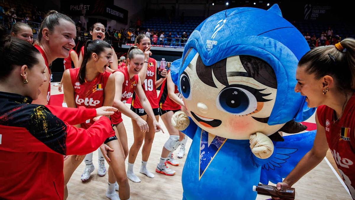 Uśmiechnięta drużyna siatkarek w czerwonych strojach świętuje z dużą, niebieską maskotką podczas meczu.