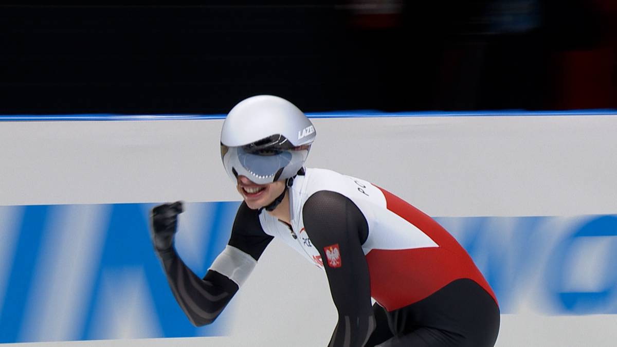 Łyżwiarz szybki w białym kasku i czerwono-białym kombinezonie unosi zaciśniętą pięść w geście triumfu.