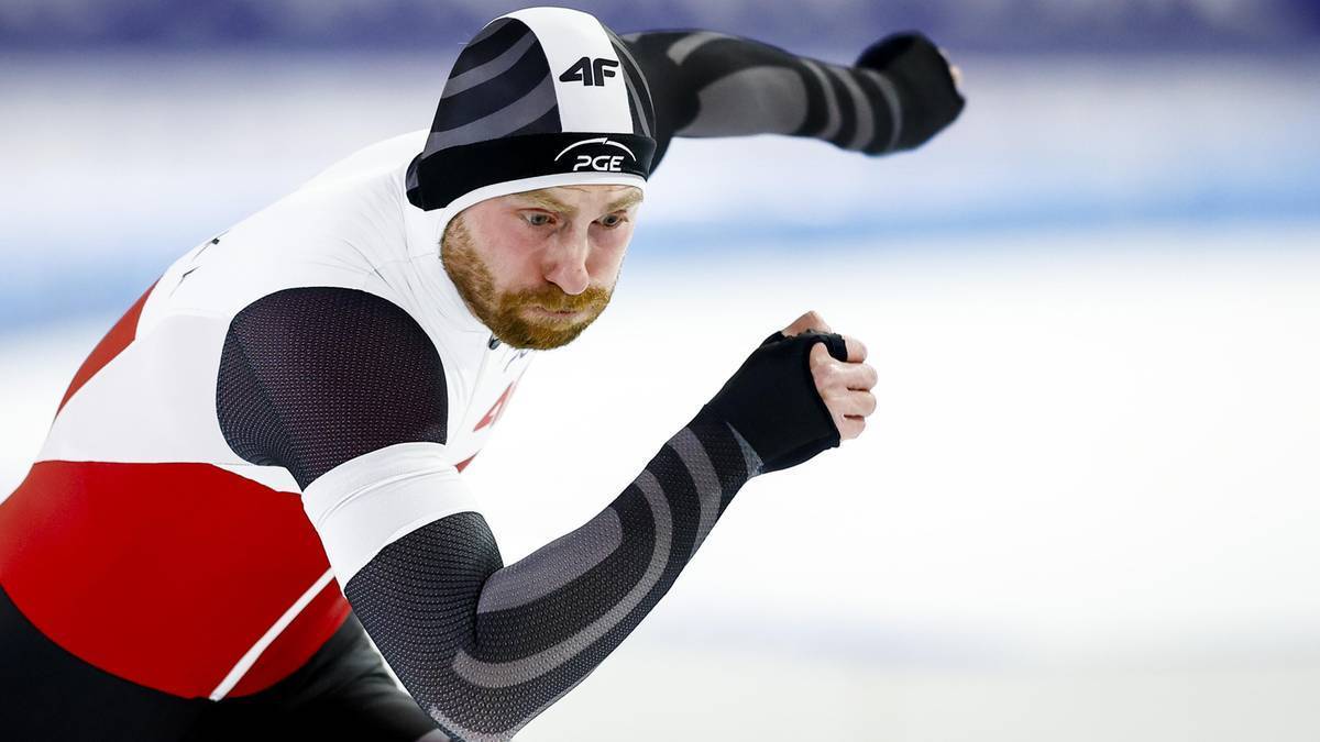 Łyżwiarz szybki w białej czapce i stroju sportowym z czerwonymi wstawkami, skupiony na torze.