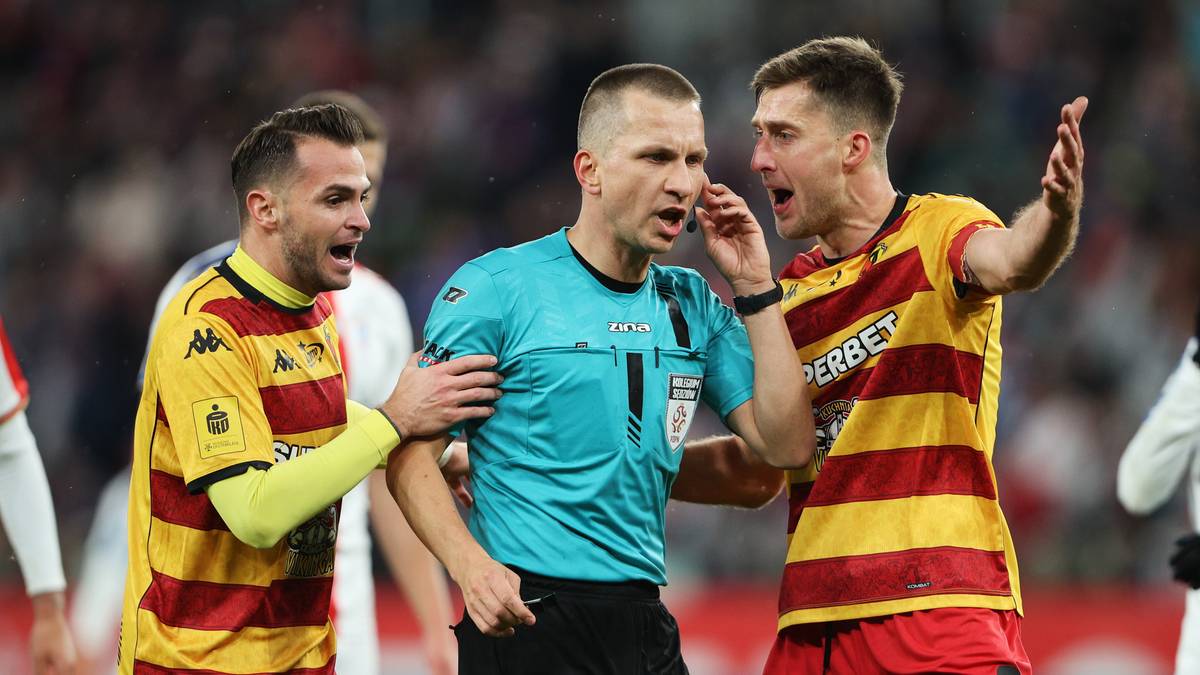 Trzech mężczyzn w strojach sportowych, dwóch w żółto-czerwonych koszulkach i jeden w niebieskim stroju sędziego, w trakcie dyskusji.