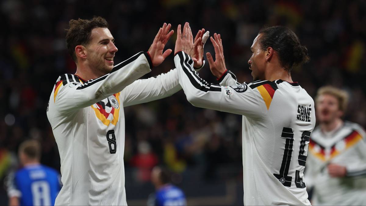 Dwóch piłkarzy reprezentacji Niemiec w białych strojach z czarnymi i czerwonymi akcentami klaszcze w dłonie, celebrując bramkę. Jeden z nich ma numer 8 na plecach, drugi numer 13. W tle widać innych graczy w tych samych strojach oraz publiczność na trybunach.