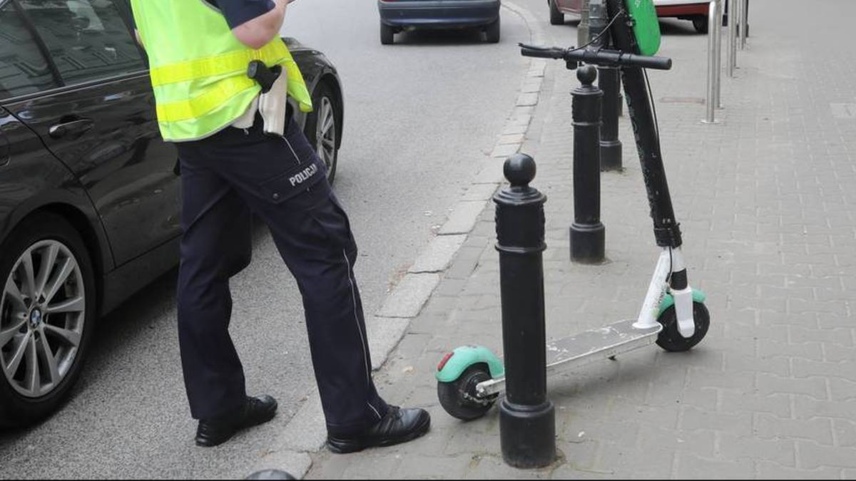 Funkcjonariusz w kamizelce odblaskowej i granatowych spodniach z napisem "POLICJA" obok zaparkowanego samochodu i hulajnogi elektrycznej.
