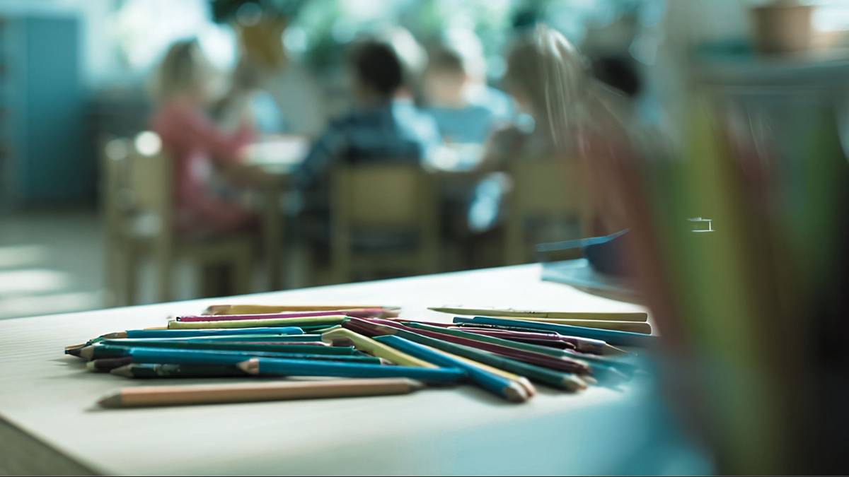 Zbliżenie na grupę kolorowych kredek leżących na stole, z rozmyciem na tle przedstawiającym dzieci w sali.