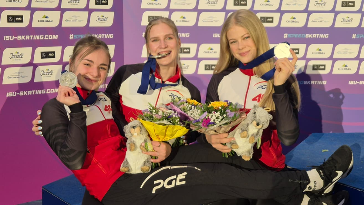 Trzy młode kobiety w strojach sportowych siedzą na podium, jedna trzyma złoty medal, druga srebrny, a trzecia prezentuje srebrny medal.
