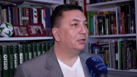 Mężczyzna w szarym garniturze i białej koszuli m&oacute;wi do mikrofonu. Za nim znajdują się p&oacute;łki z książkami i dekoracjami, w tym piłka nożna.