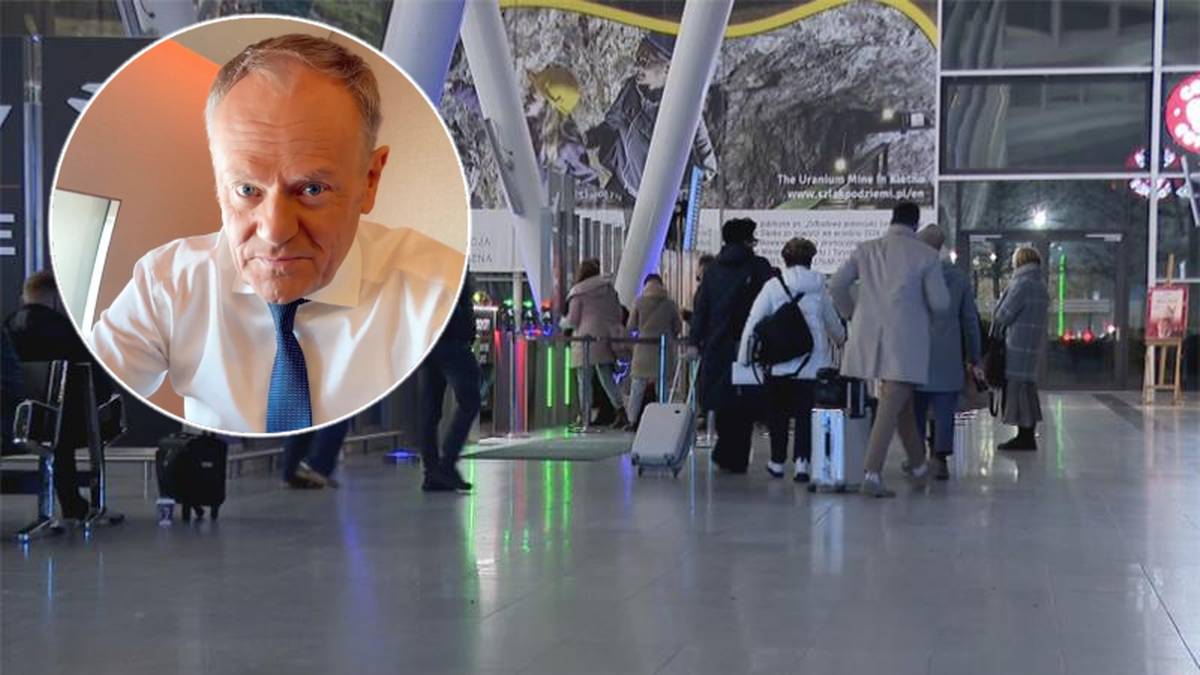 Donald Tusk przemawia na tle grupy os&oacute;b z bagażami w terminalu.