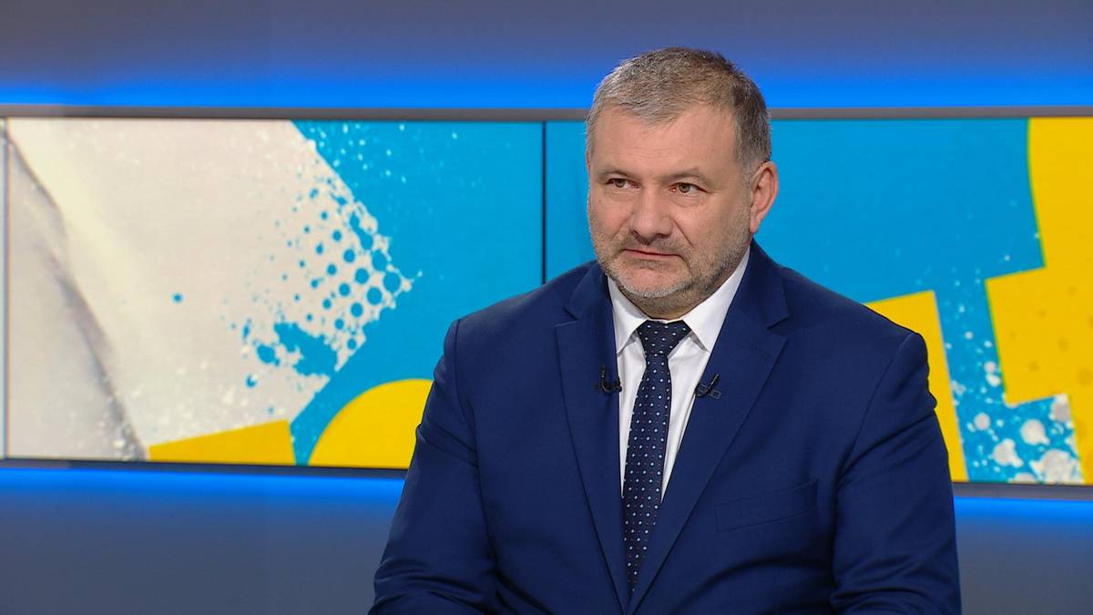 Mężczyzna w średnim wieku w granatowym garniturze i białej koszuli z krawatem, siedzi przed ekranem z kolorowym abstrakcyjnym wzorem w tle.