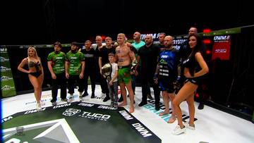 Grupa zawodnik&oacute;w MMA i ring girls pozuje do zdjęcia na macie po walce.