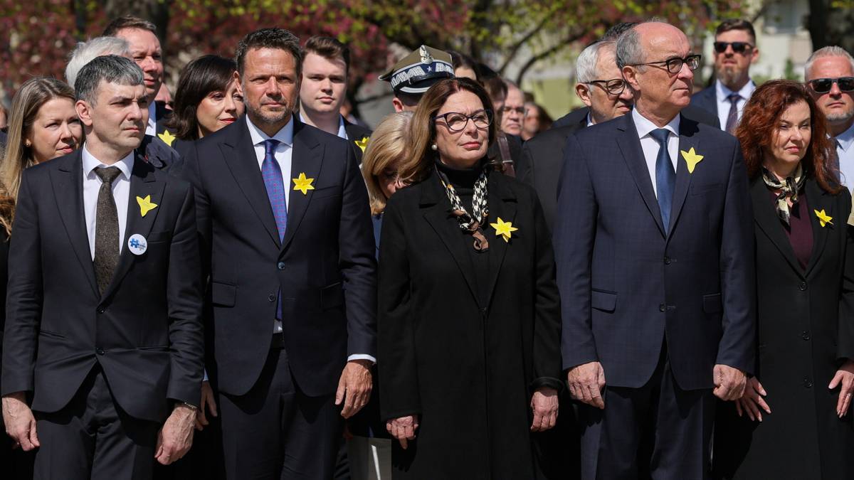 Uczestnicy uroczystości rocznicowych, w tym politycy, stojący razem, niekt&oacute;rzy z przypiętymi ż&oacute;łtymi żonkilami.