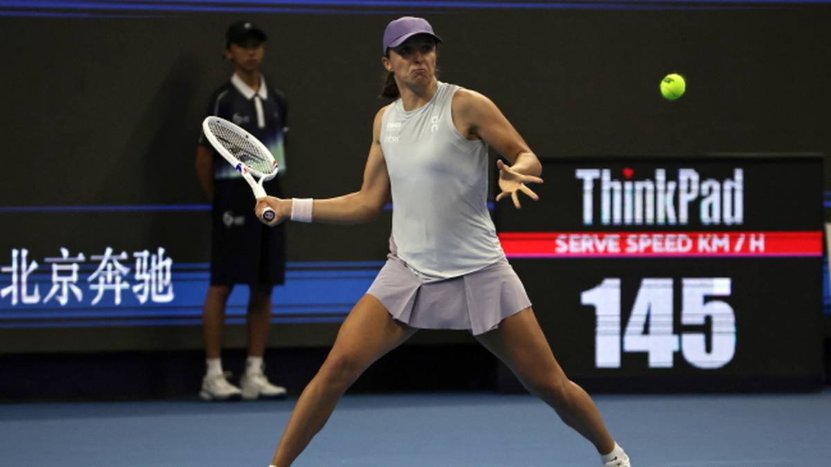 Iga Świątek podczas meczu tenisowego, w białej bluzce i fioletowej spódniczce, celuje w piłkę rakietą. Na drugim planie widać tablicę wyników.