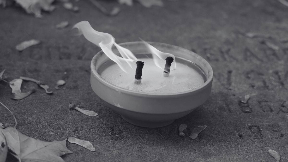 Zbliżenie na znicz palący się na nagrobku. Widać dwa płonące knoty i dym unoszący się nad płomieniami. Kamień nagrobka jest w tle, z widocznymi wyrytymi literami. Wokół leżą opadłe liście.