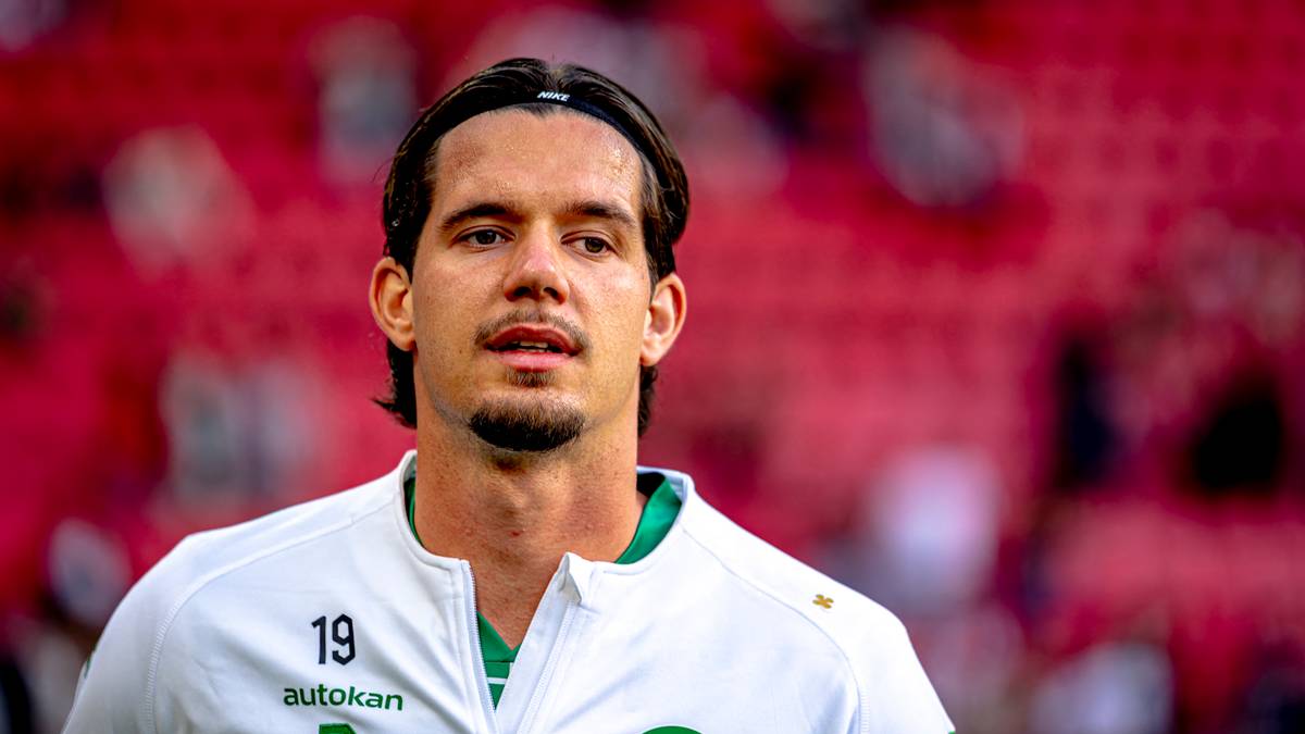 Piłkarz w białej koszulce z numerem 19 na tle czerwonych trybun.