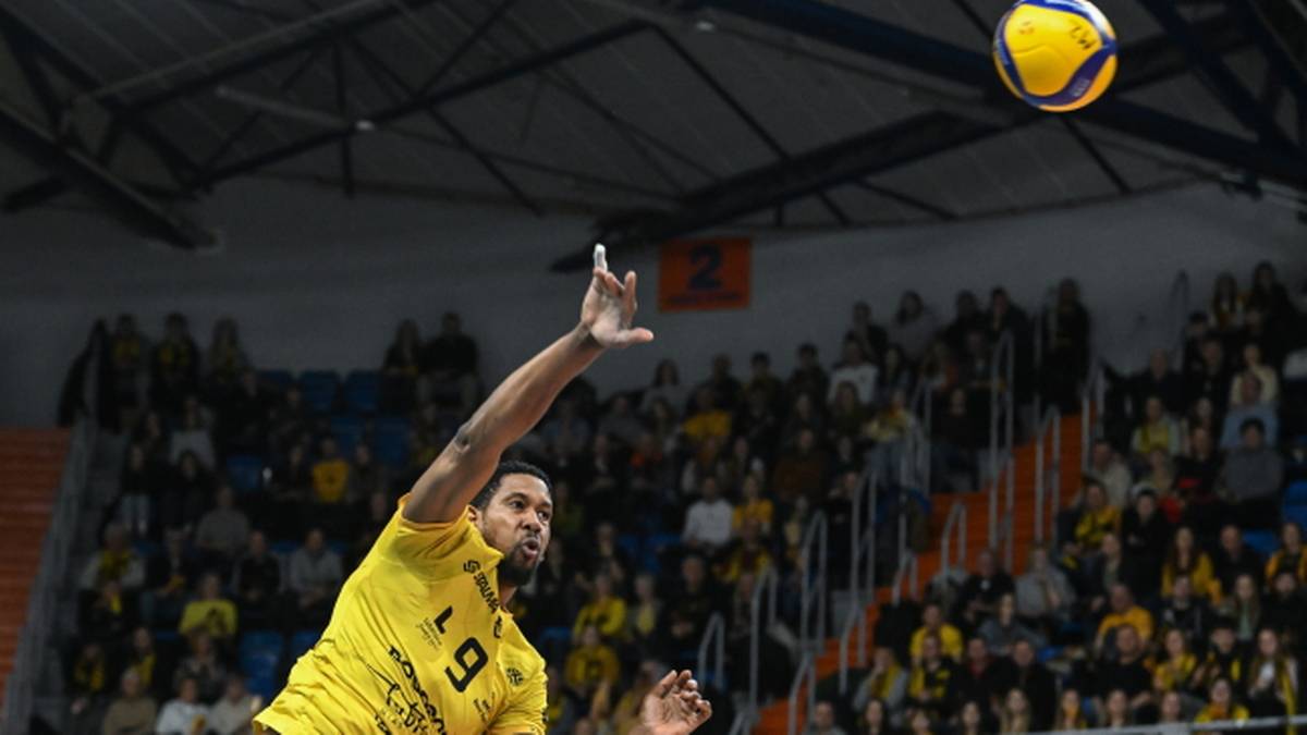 Siatkarz w żółtej koszulce wykonuje zagrywkę, w tle trybuny pełne kibiców.