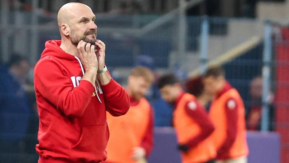 Mężczyzna w czerwonej bluzie z kapturem stoi z rękami przy twarzy, patrząc w bok. W tle widać innych mężczyzn w kamizelkach i trybuny stadionu.