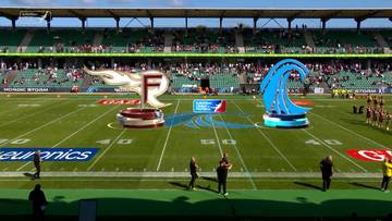 Boisko do futbolu amerykańskiego z logotypami drużyn Rhein Fire (płonące F) i Nordic Storm (niebieska fala) na podestach. W tle zielone trybuny wypełnione widzami.