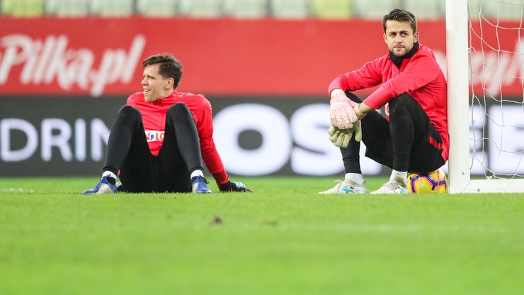 Dw&oacute;ch piłkarzy siedzi na zielonej murawie stadionu, ubranych w czerwone stroje reprezentacji Polski i rękawice bramkarskie.