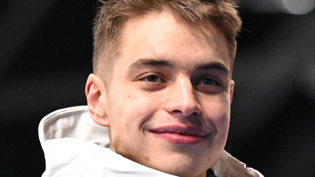 Portret młodego mężczyzny w białej kurtce sportowej, trzymającego w dłoni srebrny medal, z uśmiechem na twarzy.