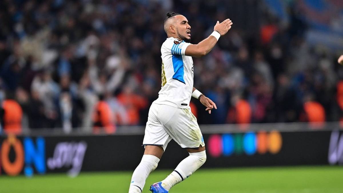 Piłkarz w białym stroju świętuje na boisku, uniesiona ręka, tłum na trybunach w tle.