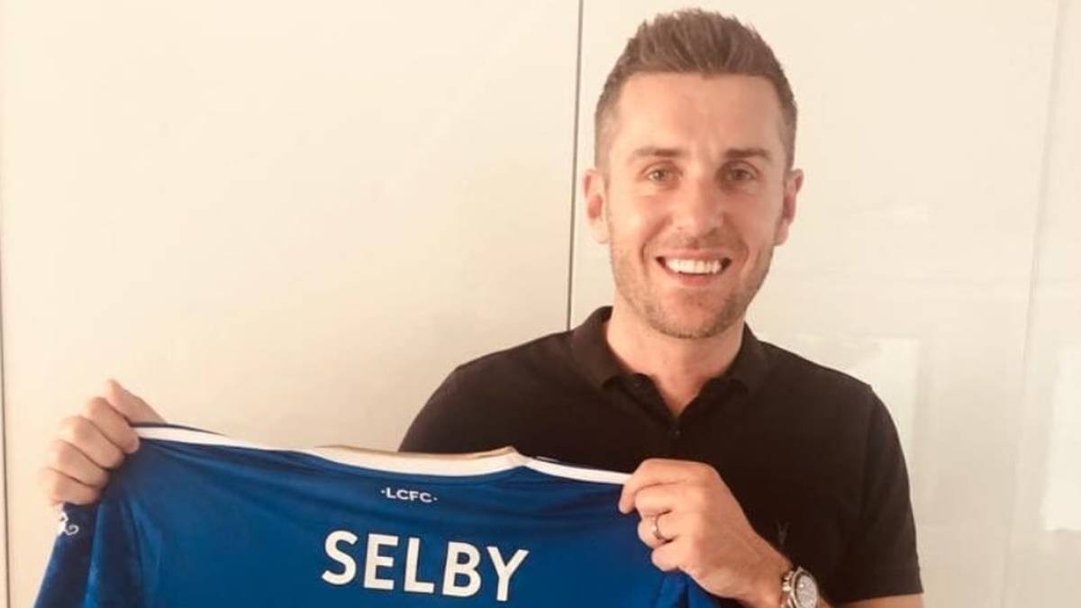 Uśmiechnięty mężczyzna trzyma niebieską koszulkę z napisem "SELBY" i numerem "4".