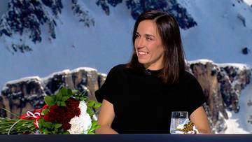 Zuzanna Witych, kobieta o ciemnych włosach, w czarnej bluzce, siedzi przed bukietem kwiat&oacute;w i szklanką wody. W tle widać zaśnieżone g&oacute;ry.