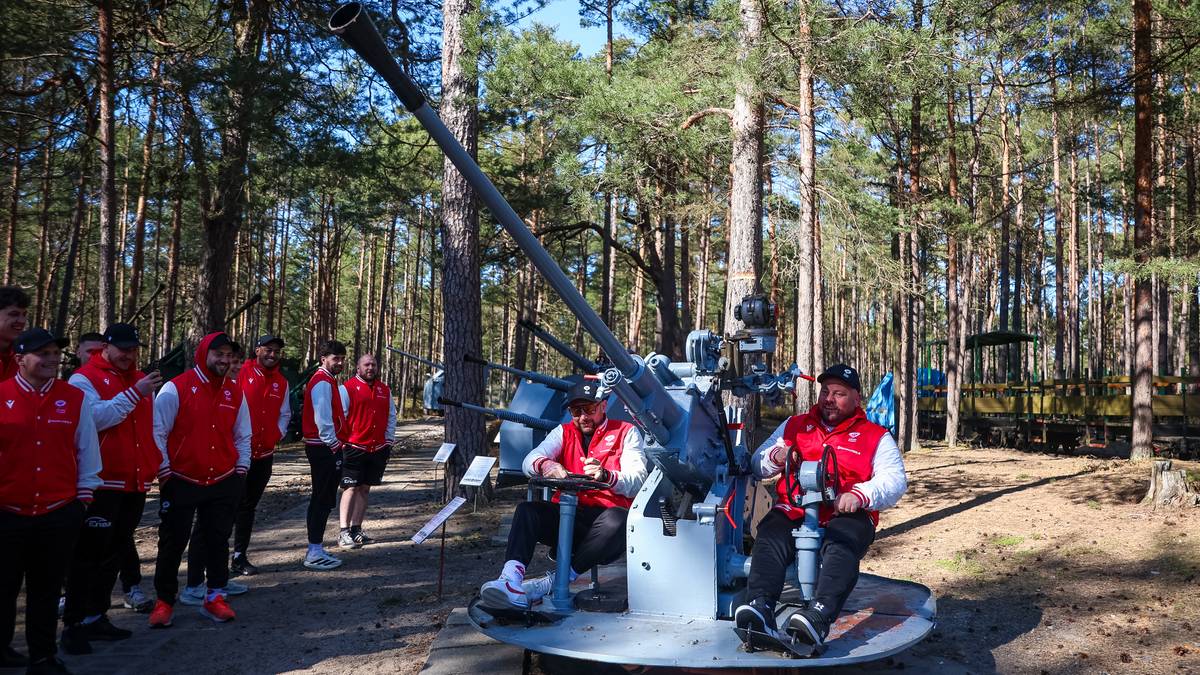 Drużyna rugby w czerwonych kamizelkach zwiedza historyczną artylerię na świeżym powietrzu, z dużą armatą w centrum uwagi.
