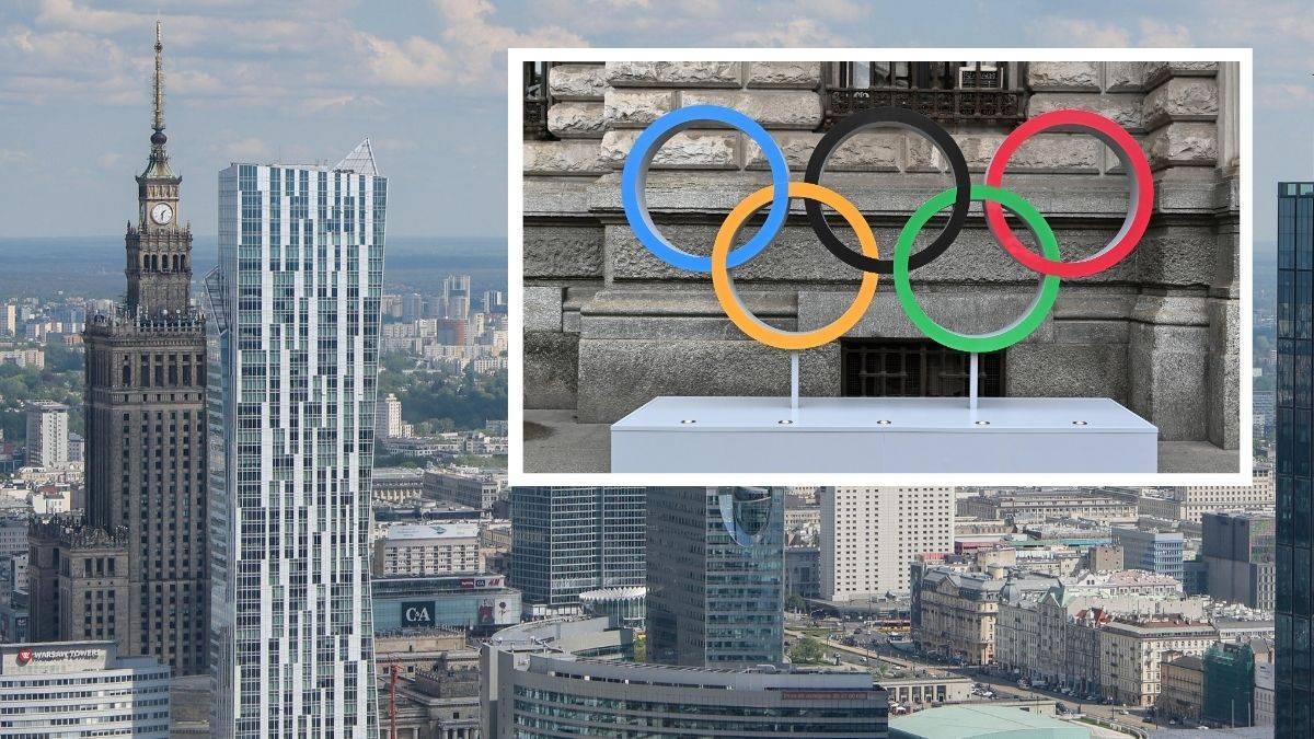 Pięć kolorowych kół olimpijskich na białym postumencie przed budynkiem, w tle panorama miejska z wieżowcami