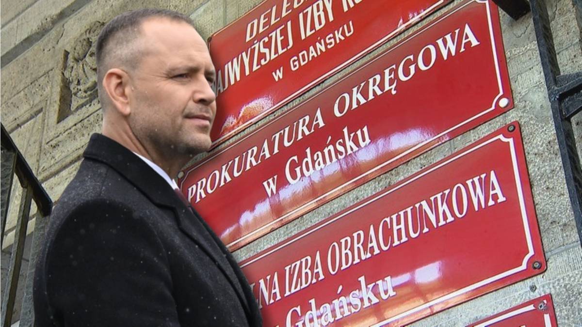 Mężczyzna w garniturze stoi przed budynkiem z czerwonymi tablicami informacyjnymi.
