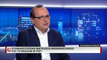 Mężczyzna w okularach i granatowej marynarce, ubrany w białą koszulę, siedzi przed ekranem z nocnym widokiem miasta. Na dole ekranu wyświetlany jest napis "STOWARZYSZENIE MATEUSZA MORAWIECKIEGO CZY TO ROZŁAM W PIS?".