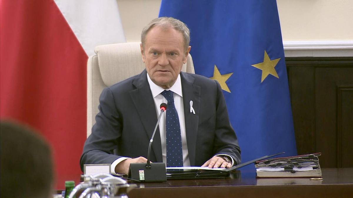 Premier Donald Tusk siedzi przy stole, za nim flagi Polski i Unii Europejskiej.