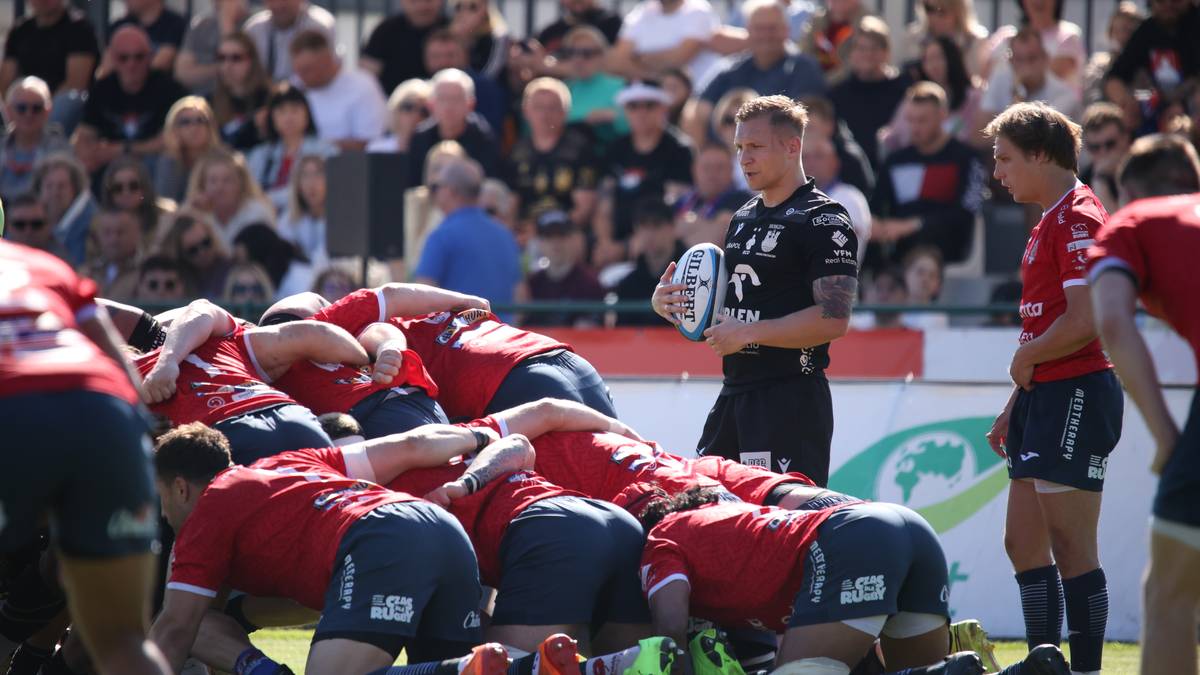 Gracze rugby w czerwonych koszulkach i spodenkach, z widoczną grupą na pierwszym planie, grają w meczu. Jeden z nich trzyma piłkę do rugby. W tle widać publiczność na trybunach.