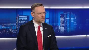 Mężczyzna w garniturze i krawacie siedzi w studiu telewizyjnym na tle ekran&oacute;w z nocną panoramą miasta.