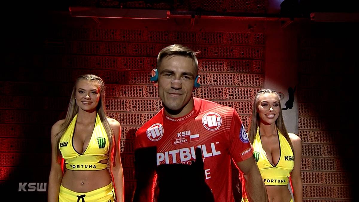 Uśmiechnięty wojownik MMA w czerwonej koszulce Pitbull, otoczony przez dwie hostessy w żółtych strojach, na tle ceglanej ściany i oświetlenia scenicznego.