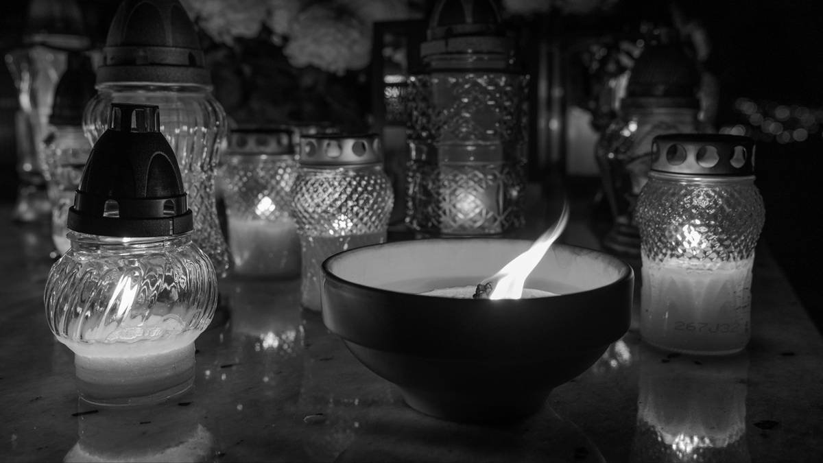 Zbliżenie na płonący znicz na tle innych zniczy i kwiat&oacute;w.