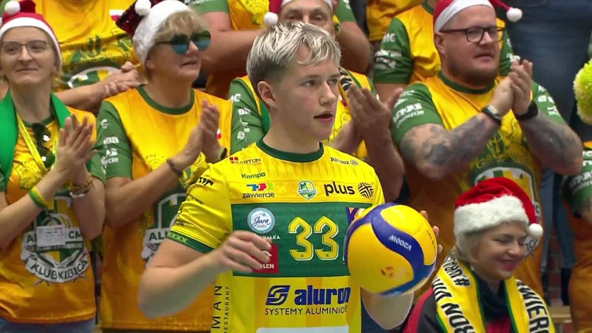 Młody siatkarz w żółtej koszulce z numerem 33 trzyma piłkę siatkową, otoczony przez kibiców w świątecznych czapkach.