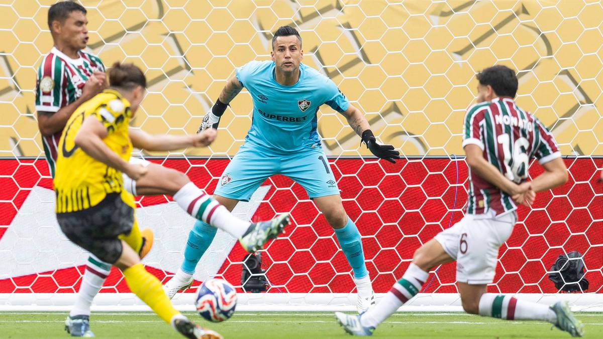 Dwóch piłkarzy w biało-zielonych strojach próbuje odebrać piłkę piłkarzowi w żółtej koszulce. Bramkarz w niebieskim stroju obserwuje sytuację.