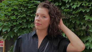 Młoda kobieta o kręconych, ciemnych włosach, ubrana w czarną koszulę, patrzy w obiektyw na tle zielonych liści.