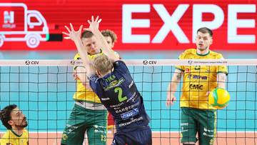 Siatkarze walczą o piłkę przy siatce, jeden z nich w granatowym stroju z numerem 2 pr&oacute;buje zablokować atak.