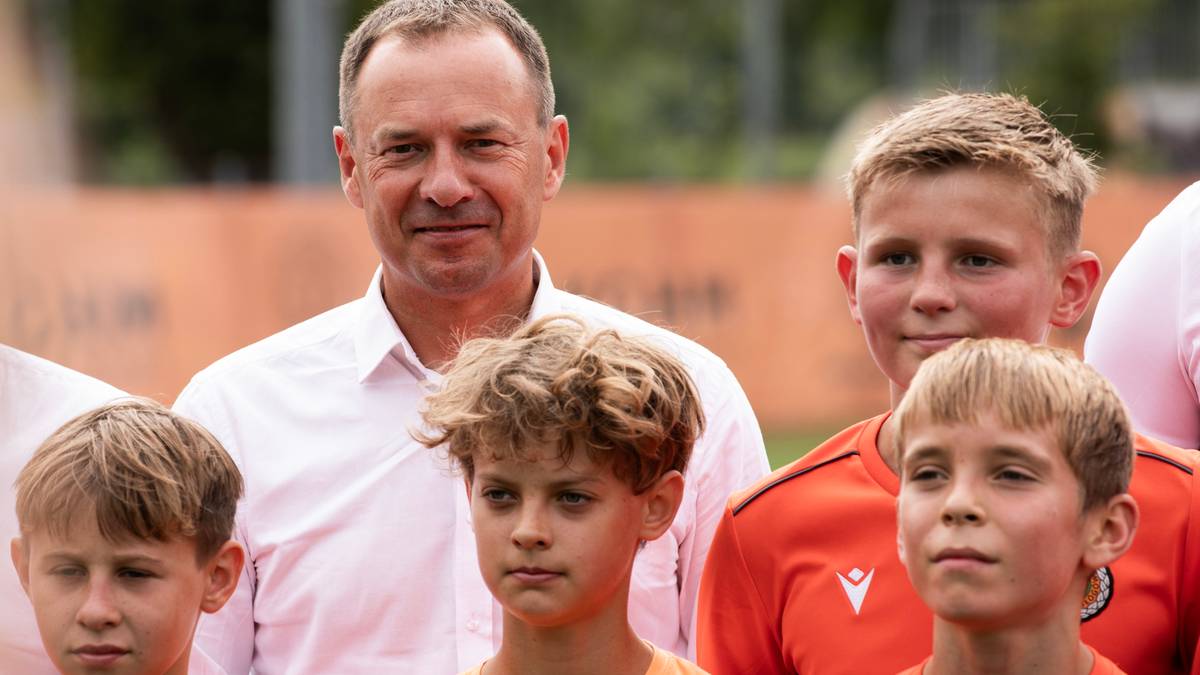 Mężczyzna w białej koszuli i trzej chłopcy w pomarańczowych strojach sportowych stoją na zewnątrz, patrząc w kierunku obiektywu.