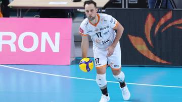 Siatkarz w białej koszulce z numerem 17 i białych spodenkach prowadzi piłkę do siatkówki na boisku.